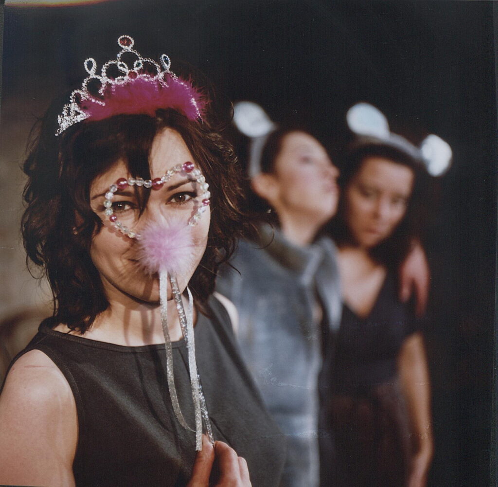 Zdjęcie ze spektaklu Wszystko o kobietach. Zbliżenie. Aktorka w zabawkowej koronie na głowie. Do twarzy przykłada różdżkę w kształcie serca. W tle dwie aktorki obejmują się, na głowach mają opaski z króliczymi uszami.