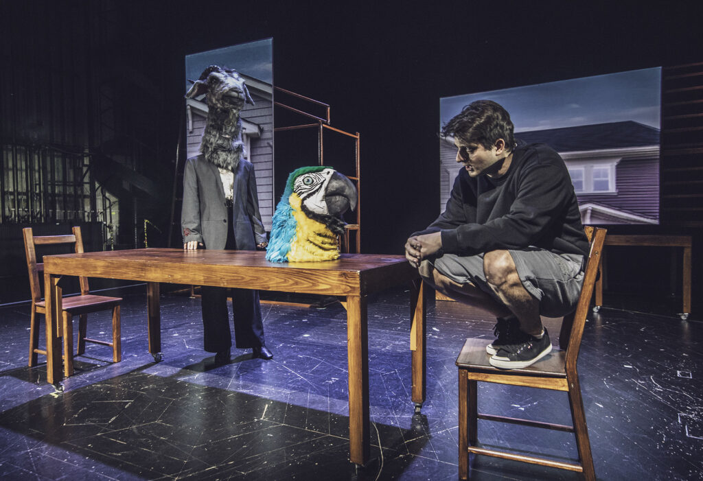 Zdjęcie ze spektaklu Kruk z Tower. Młody aktor kuca na krześle przy stole. Po drugiej stronie stoi kobieta z głową kozła. Na stole stoi głowa papugi. Za nimi w tle są wyświetlone projekcje domu.