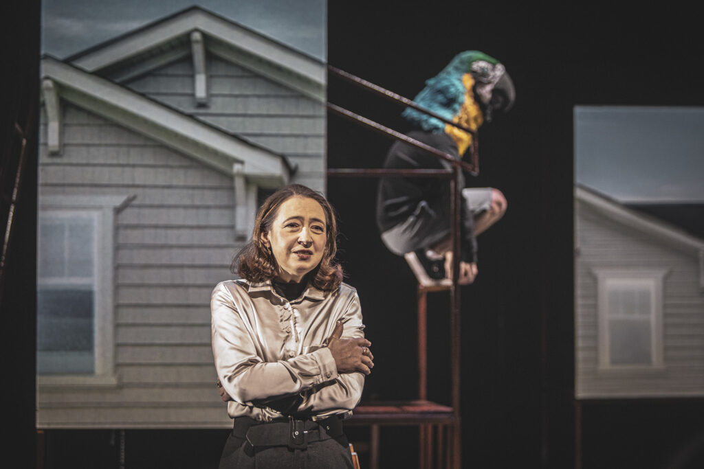Zdjęcie ze spektaklu Kruk z Tower. Półzbliżenie. Na pierwszym planie stoi aktorka w średnim wieku. Ma skrzyżowane ręce. Z tyłu aktor z głową papugi siedzi na stelażowej konstrukcji między ekranami z projekcją domu.