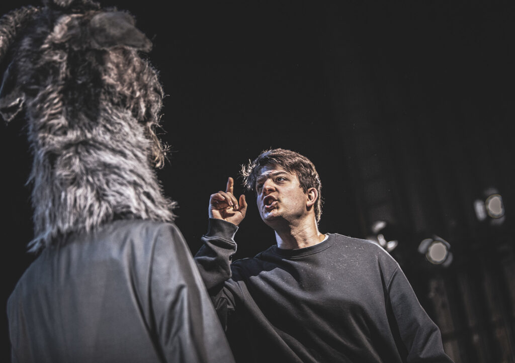 Zdjęcie ze spektaklu Kruk z Tower. Młody aktor i aktorka z głową kozła stoją naprzeciwko siebie. Aktor otwiera usta, podnosi palec wskazujący, jest zdenerwowany.