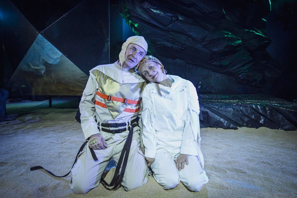 Zdjęcie ze spektaklu Mały Książę. Aktor i aktorka w białych kombinezonach i czapkach pilotkach klęczą na piasku. Opierają się o siebie. W tle jest czarna skała i lustrzany kryształ.