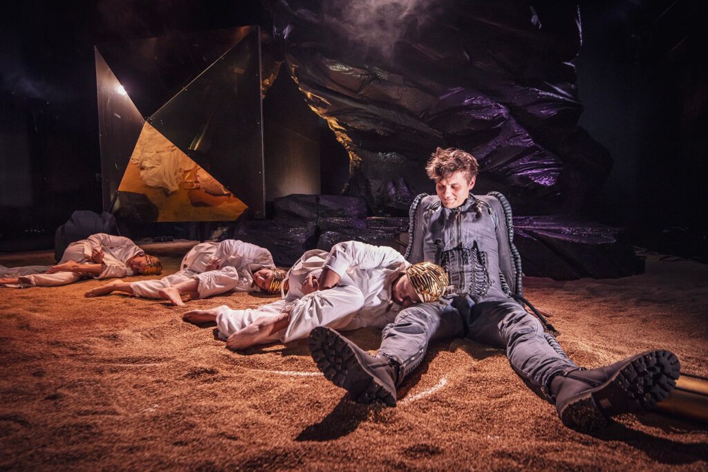 Zdjęcie ze spektaklu Mały Książę. Aktor w ciemnym kombinezonie siedzi na piasku. Aktorka w białym kombinezonie leży na piasku, głowę opiera na nodze aktora. Za nią leżą dwie aktorki w kombinezonach. W tle jest czarna skała i lustrzany kryształ.