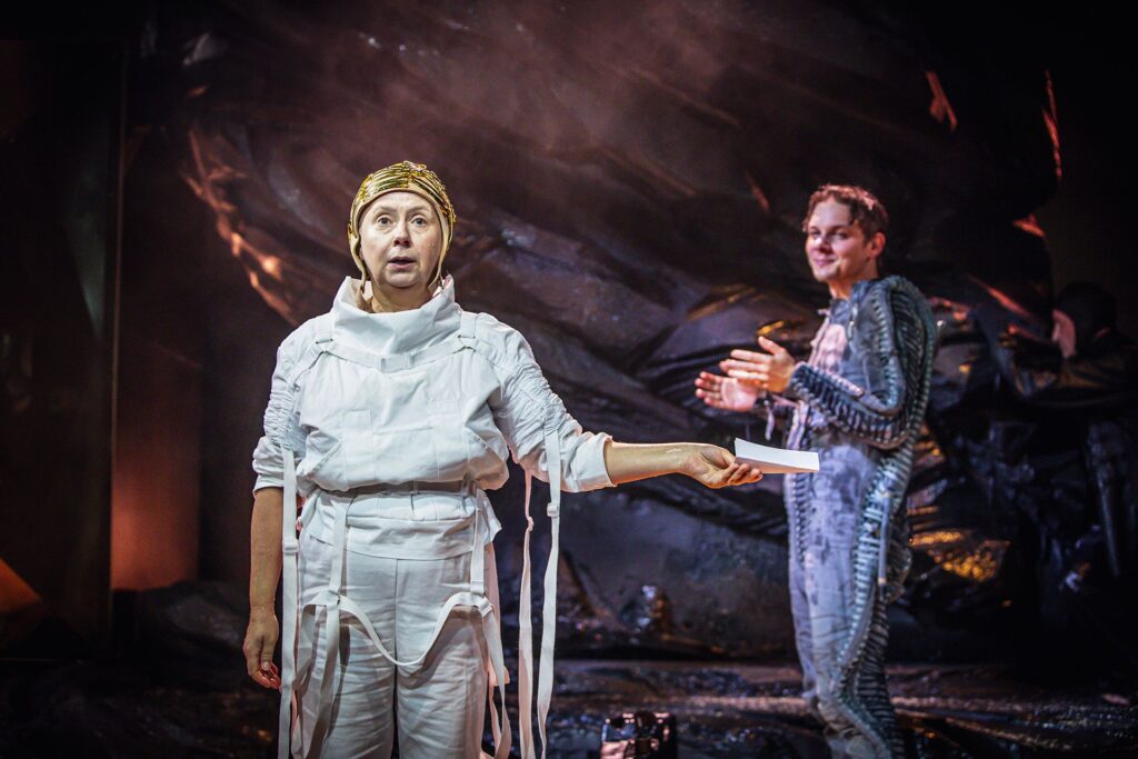 Zdjęcie ze spektaklu Mały Książę. Aktorka w białym kombinezonie i czapce pilotce trzyma w dłoni kartkę papieru. Za nią stoi aktor w ciemnym kombinezonie, patrzy na aktorkę i się uśmiecha. W tle jest czarna skała.