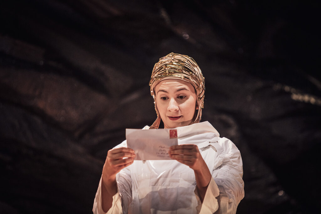 Zdjęcie ze spektaklu Mały Książę. Zbliżenie. Młoda aktorka w białym kombinezonie trzyma w dłoniach zaadresowaną kopertę.