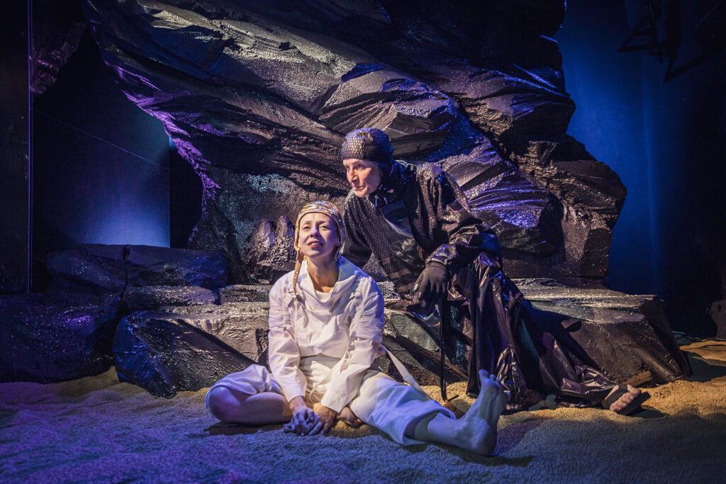 Zdjęcie ze spektaklu Mały Książę. Aktorka w białym kombinezonie i czapce pilotce siedzi na piasku. Nad nią pochyla się aktor w stroju węża. Ma czapkę i długi czarny płaszcz w łuski. W tle jest czarna skała.