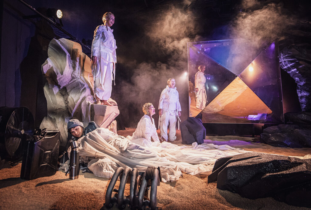 Zdjęcie ze spektaklu Mały Książę. Aktor w ciemnym kombinezonie leży na piasku przykryty długim materiałem. Z tyłu są trzy aktorki w białych kombinezonach. Jedna aktorka stoi na skale, druga klęczy na piasku, a trzecia stoi obok lustrzanego kryształu.