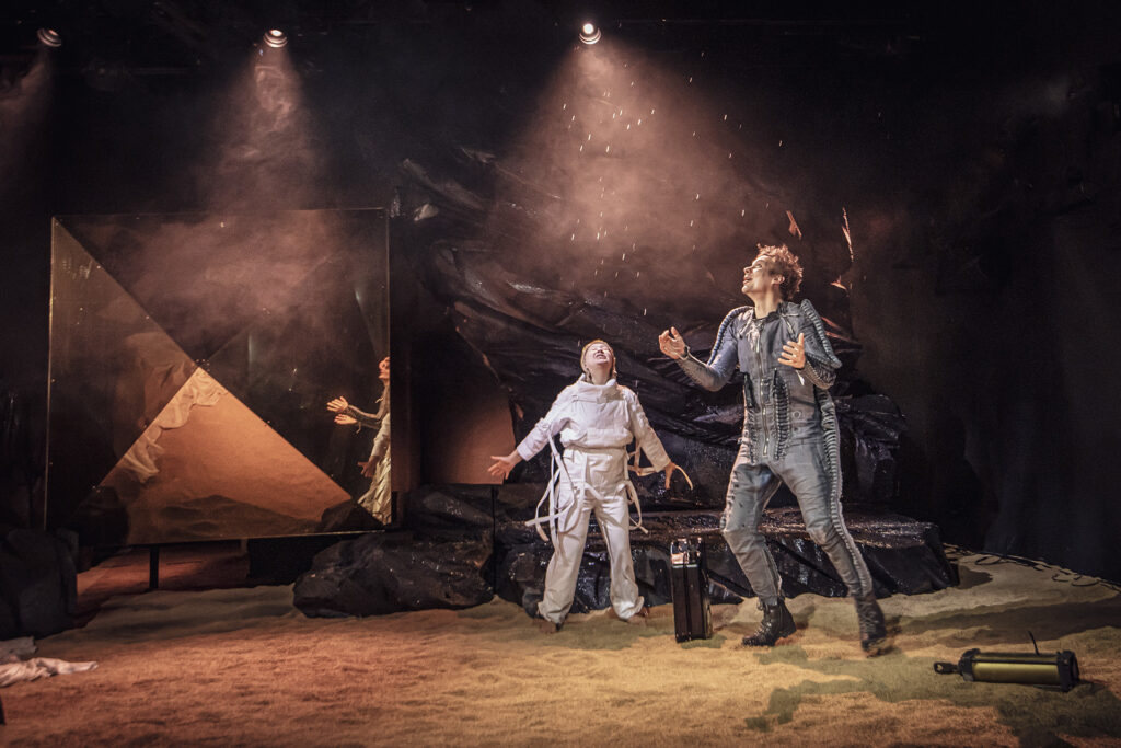 Zdjęcie ze spektaklu Mały Książę. Aktorka w białym kombinezonie i aktor w ciemnym kombinezonie są w ruchu. Rozkładają ręce i patrzą do góry. Z sufitu spadają na nich krople wody. W tle jest czarna skała i lustrzany kryształ.