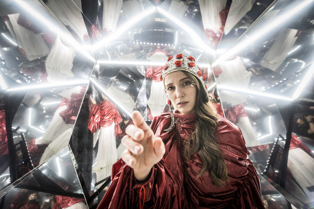 djęcie ze spektaklu Mały Książę. Aktorka przebrana za różę. Ma czerwony strój, czapkę pilotkę z naszytymi małymi różami. Wyciąga rękę do przodu. Wokół niej jest wiele luster.