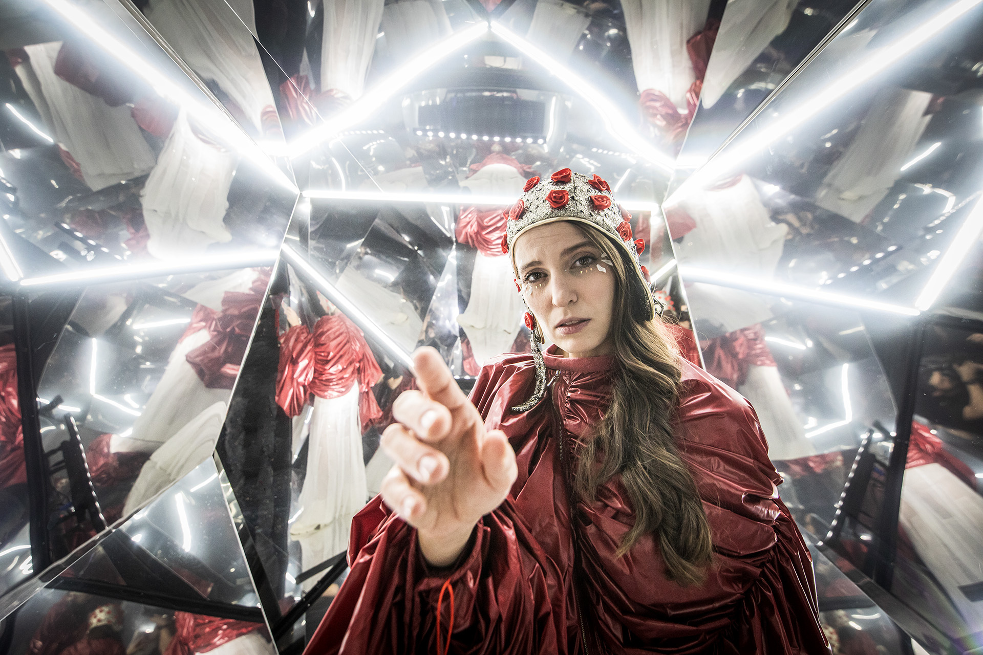 djęcie ze spektaklu Mały Książę. Aktorka przebrana za różę. Ma czerwony strój, czapkę pilotkę z naszytymi małymi różami. Wyciąga rękę do przodu. Wokół niej jest wiele luster.