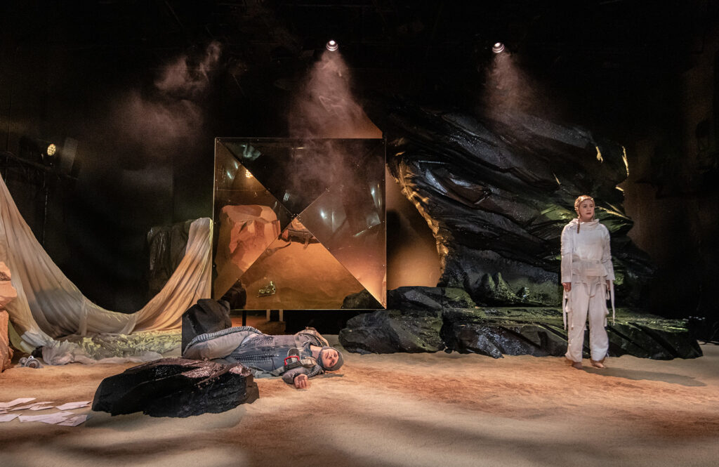 Zdjęcie ze spektaklu Mały Książę. Aktor w ciemnym kombinezonie leży na piasku. Obok stoi aktorka w białym kombinezonie. Mają smutne miny. W tle jest czarna skała i lustrzany kryształ.