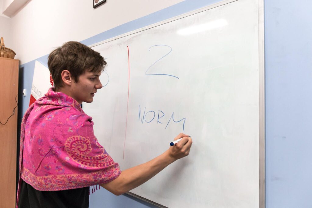 djęcie ze spektaklu Trik Patryka. Aktor w chuście na ramionach pisze markerem na tablicy magnetycznej litery NORM.