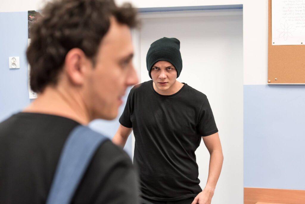 Zdjęcie ze spektaklu Trik Patryka. Zbliżenie. Na pierwszym planie aktor stoi tyłem i patrzy w bok. W jego stronę zmierza drugi aktor w czapce. Aktor w czapce ma poważną minę. Są w klasie szkolnej.