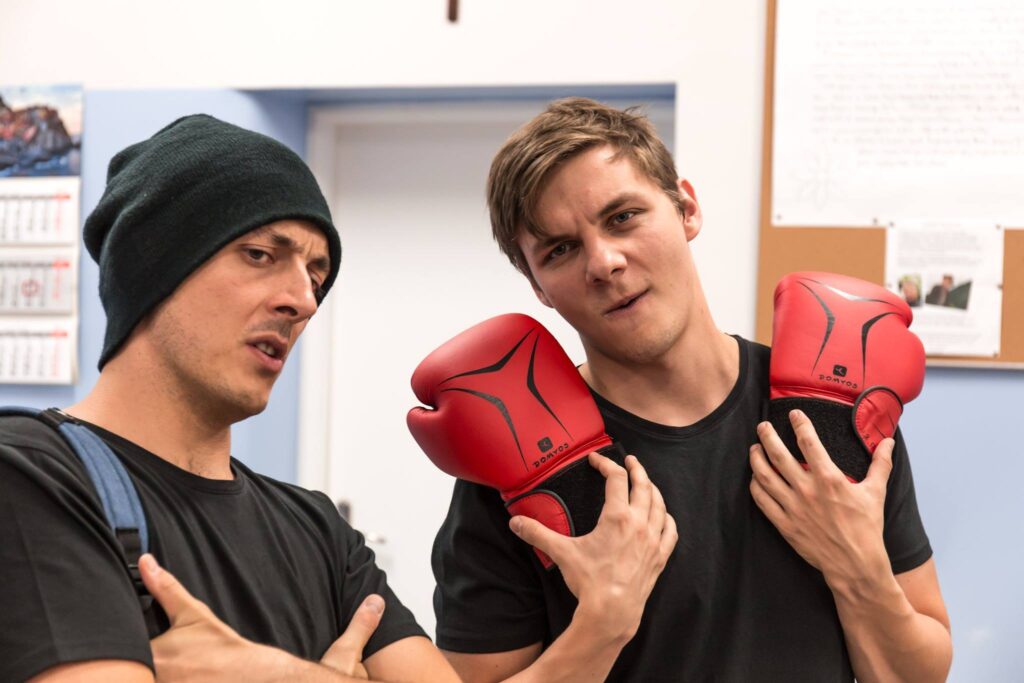 Zdjęcie ze spektaklu Trik Patryka. Zbliżenie. Dwaj młodzi aktorzy. Jeden ma czapkę, krzyżuje ręce i trzyma dłonie pod pachami. Drugi trzyma rękawice bokserskie. Mają groźne miny. Są w klasie szkolnej.