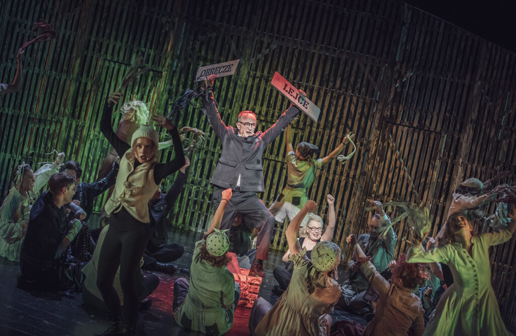 Zdjęcie ze spektaklu Folwark zwierzęcy. Na środku na drewnianej skrzynce stoi aktor w szarym stroju. Podnosi ręce. W rękach trzyma dwie tabliczki z napisami Obręcze i Lejce. Wokół niego tańczą pozostali aktorzy i aktorki.