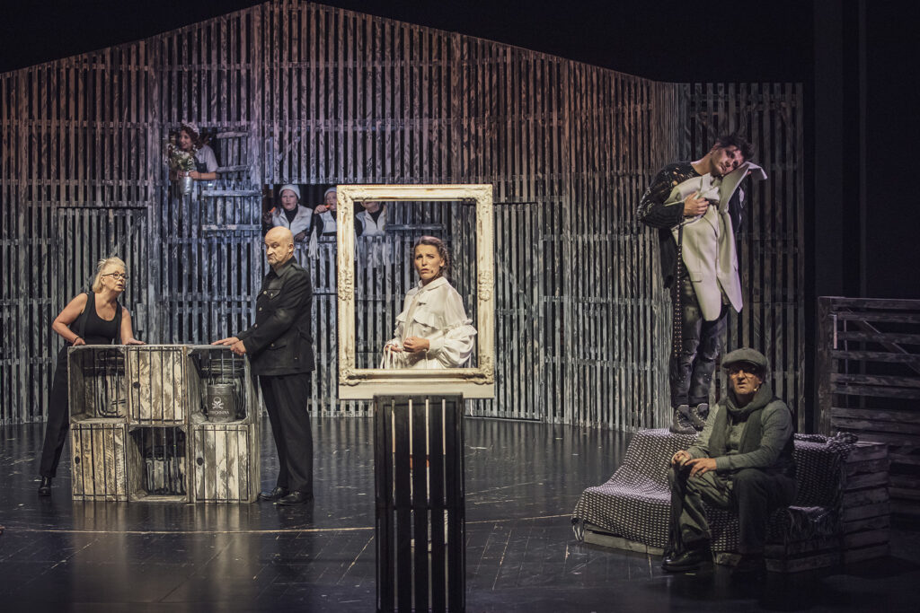 Zdjęcie ze spektaklu Folwark zwierzęcy. Na środku za pustą ramą obrazu stoi aktorka. Po lewej aktorzy stoją przy drewnianych skrzynkach. Po prawej na kanapie jeden aktor siedzi, a drugi stoi i przytula się do marynarki. W tle w oknach stodoły.