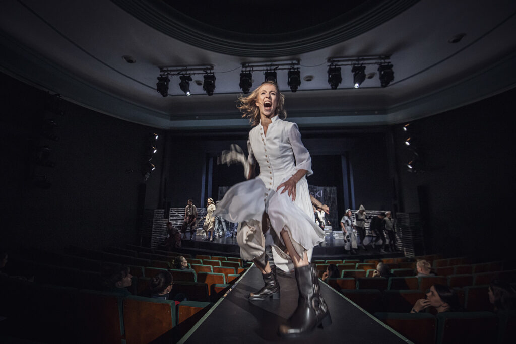 Zdjęcie ze spektaklu Folwark zwierzęcy. Aktorka w roli Gęsi tańczy na rampie. Zdjęcie w ruchu. W tle jest widownia i scena, na której są pozostali aktorzy.