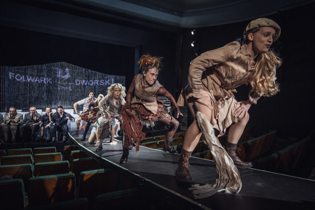 Zdjęcie ze spektaklu Folwark zwierzęcy. Aktorzy i aktorki tańczą na rampie. Za nimi na brzegu sceny siedzą pozostali aktorzy i aktorki. W tle na ścianie stodoły jest napis Folwark Dworski.