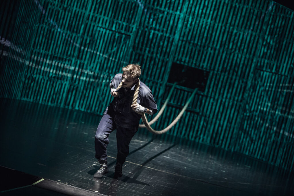 Zdjęcie ze spektaklu Folwark zwierzęcy. Aktor ciągnie długi sznur na ramionach.