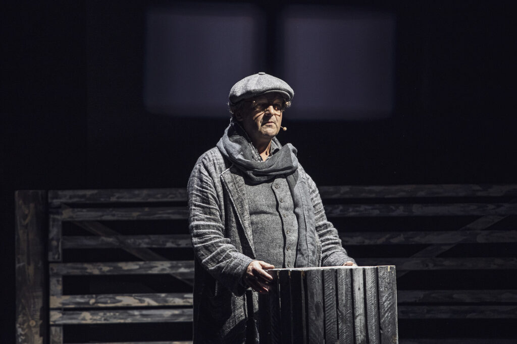 Zdjęcie ze spektaklu Folwark zwierzęcy. Aktor w szarym stroju i kaszkiecie trzyma w dłoniach drewnianą skrzynkę.