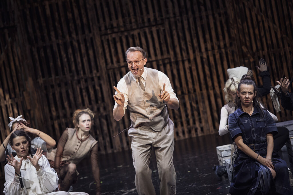 Zdjęcie ze spektaklu Folwark zwierzęcy. Na środku stoi aktor w kamizelce, otwiera usta i unosi palce wskazujące. Wokół niego na drewnianych skrzynkach siedzą aktorki, mają niezadowolone miny.