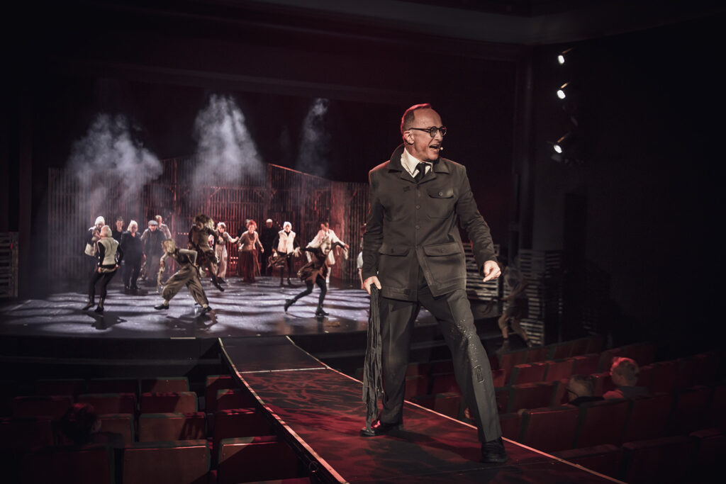 Zdjęcie ze spektaklu Folwark zwierzęcy. Na rampie stoi aktor w czarnym stroju z pejczem symbolizującym ogon w dłoni. Za nim na scenie pozostali aktorzy i aktorki biegają w koło.