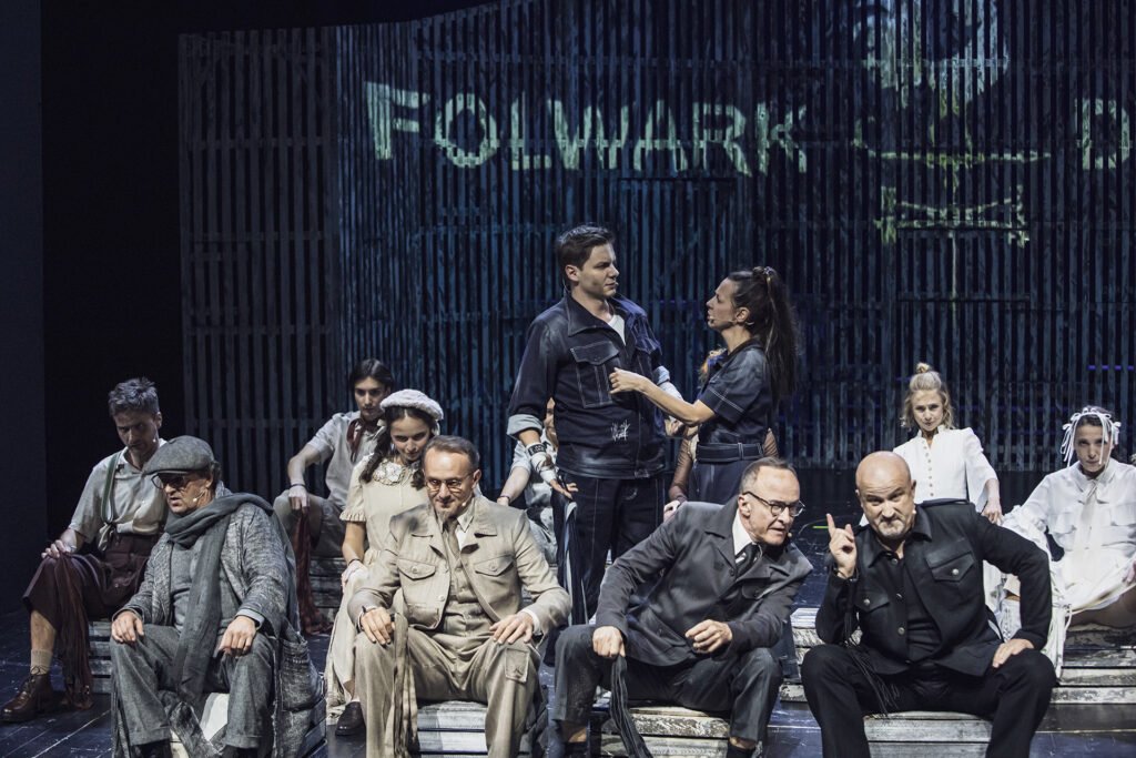 Zdjęcie ze spektaklu Folwark zwierzęcy. Na środku aktor i aktorka w jeansowych strojach stoją i patrzą na siebie. Wokół nich siedzą pozostali aktorzy i aktorki. W tle na ścianie stodoły jest napis Folwark.