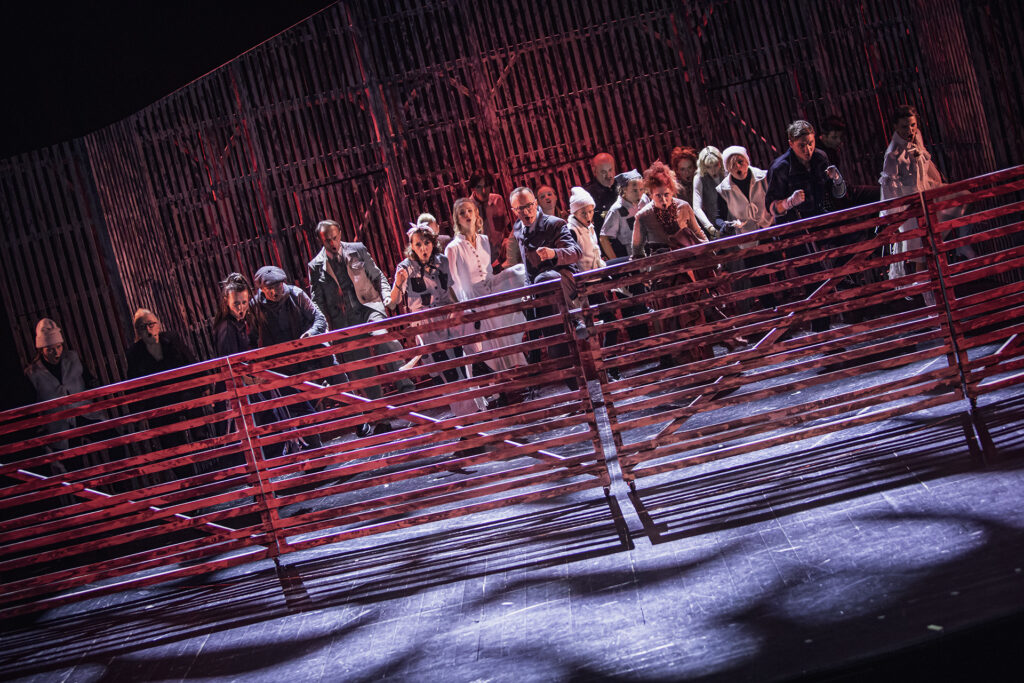 Zdjęcie ze spektaklu Folwark zwierzęcy. Zdjęcie zabarwione na czerwono. Scena grupowa. Aktorzy i aktorki stoją za ogrodzeniem.