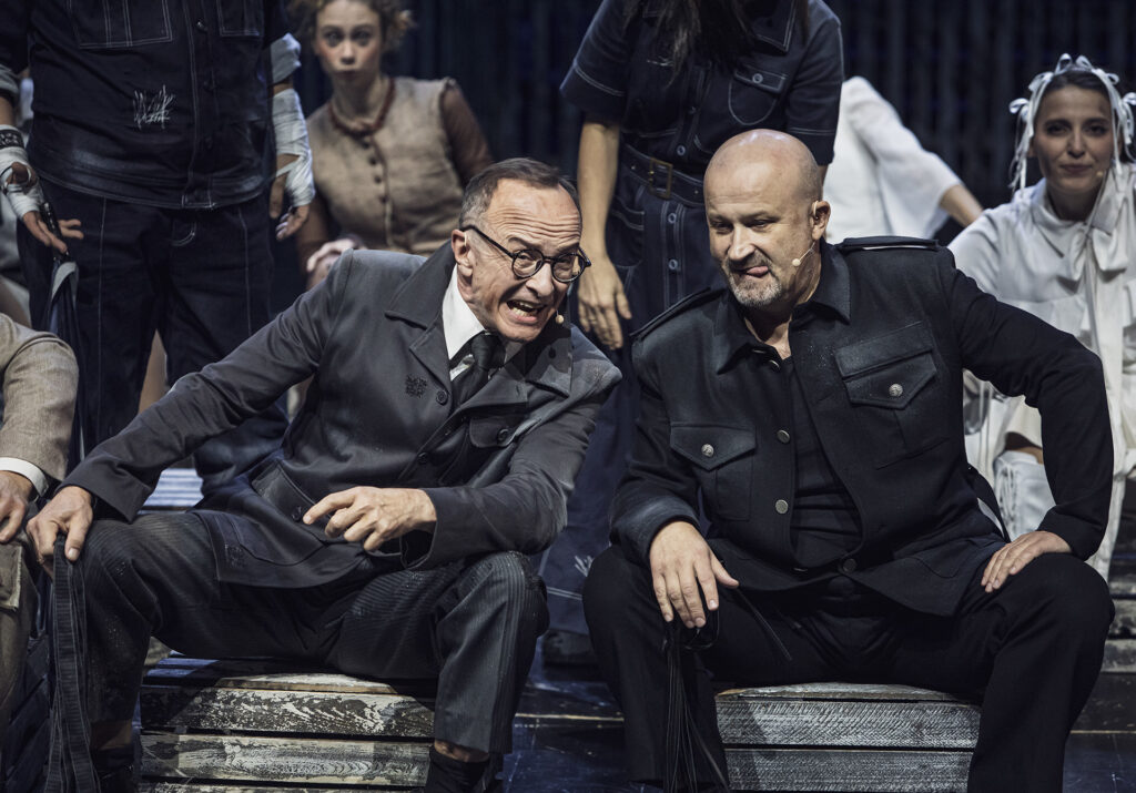 Zdjęcie ze spektaklu Folwark zwierzęcy. Na pierwszym planie dwóch aktorów w szarych strojach siedzi na drewnianych skrzynkach i nachyla się do siebie. Za nimi siedzą pozostali aktorzy i aktorki.