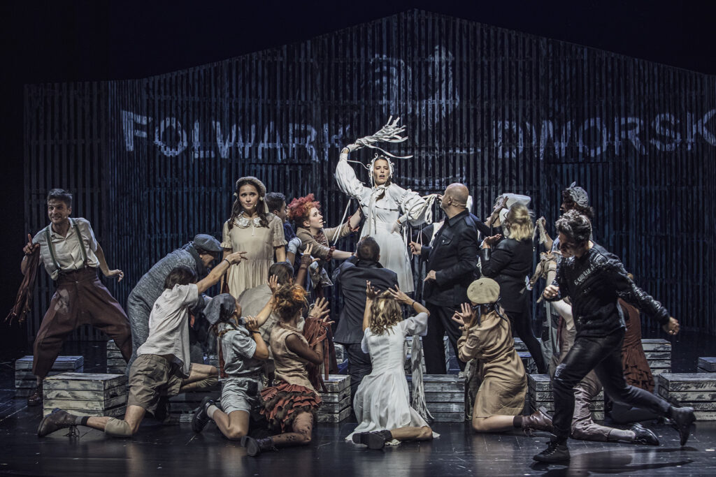 Zdjęcie ze spektaklu Folwark zwierzęcy. Scena grupowa. Na środku stoi aktorka w roli Gęsi. Wokół niej są pozostali aktorzy i aktorki, stoją lub klęczą. Prawie wszyscy patrzą na nią. W tle na ścianie stodoły jest napis Folwark Dworski.