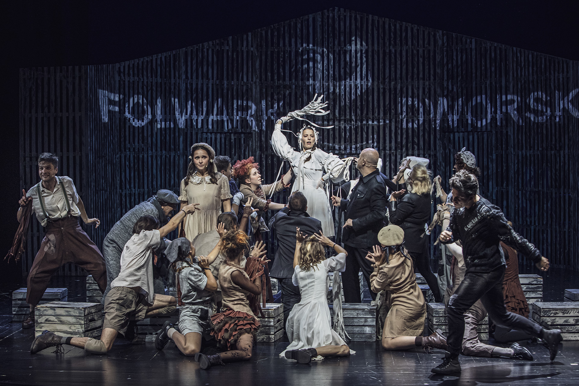 Zdjęcie ze spektaklu Folwark zwierzęcy. Scena grupowa. Na środku stoi aktorka w roli Gęsi. Wokół niej są pozostali aktorzy i aktorki, stoją lub klęczą. Prawie wszyscy patrzą na nią. W tle na ścianie stodoły jest napis Folwark Dworski.