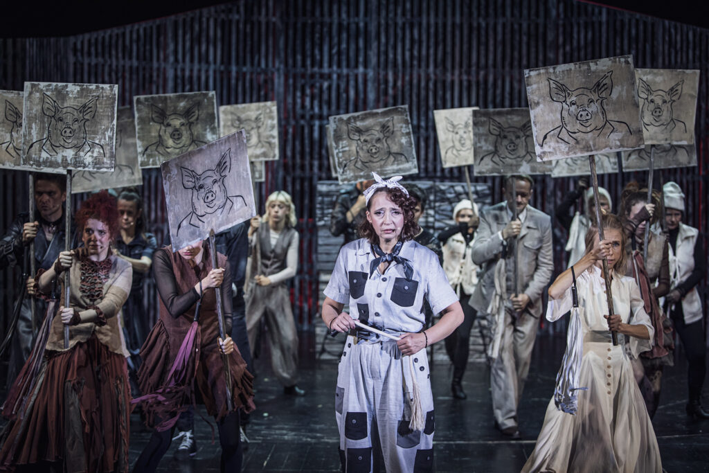 Zdjęcie ze spektaklu Folwark zwierzęcy. Na środku stoi aktorka w roli Krowy z pejczem symbolizującym ogon w dłoni. Wokół niej stoją pozostali aktorzy i aktorki. W dłoniach trzymają tabliczki na kijach. Na każdej tabliczce narysowana jest świnia.