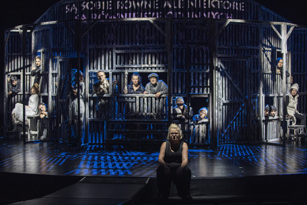 Zdjęcie ze spektaklu Folwark zwierzęcy. Zdjęcie zabarwione na niebiesko. Na brzegu sceny siedzi aktorka. Za nią w oknach stodoły są pozostali aktorzy i aktorki. Na stodole wyświetla się napis „(…) są sobie równie, ale niektóre są równiejsze od innych”.