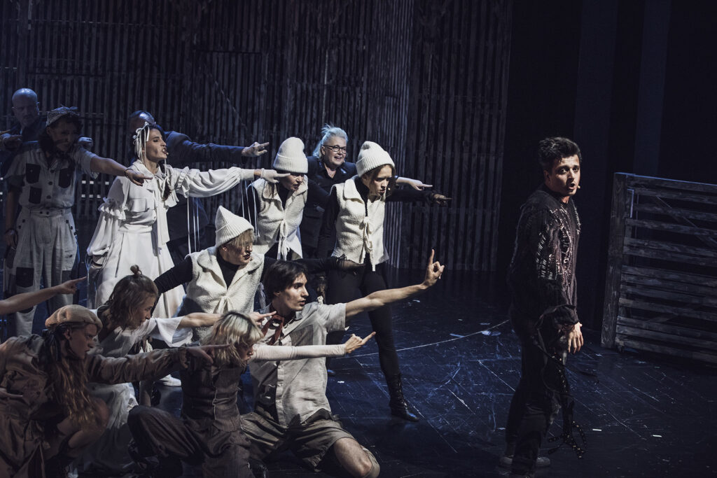 Zdjęcie ze spektaklu Folwark zwierzęcy. Aktor w czarnym stroju stoi z przodu. Za nim są aktorzy i aktorki w jasnych strojach. Wszyscy wskazują palcem na aktora z przodu.