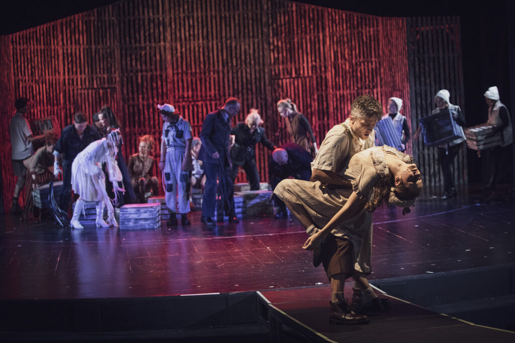 Zdjęcie ze spektaklu Folwark zwierzęcy. Scena zabarwiona na czerwono. Na pierwszym planie aktorka i aktor obejmują się. W tle pozostali aktorzy i aktorki układają drewniane skrzynki.