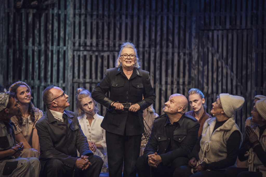 Zdjęcie ze spektaklu Folwark zwierzęcy. Na środku stoi aktorka w czarnym stroju i okularach, uśmiecha się. Wokół niej siedzą pozostali aktorzy i aktorki. Jedni się uśmiechają, inni mają zmartwione miny.