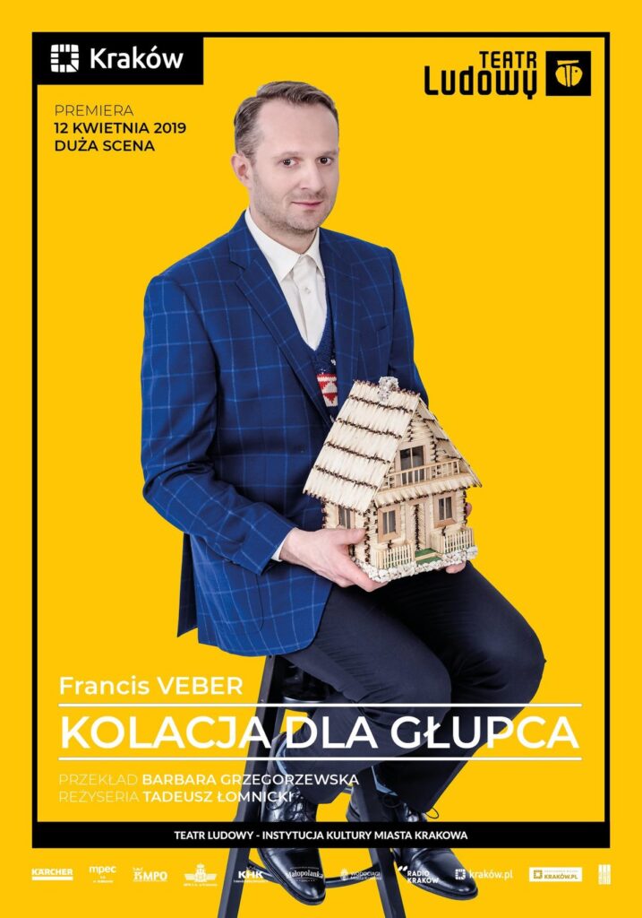 Plakat do spektaklu Kolacja dla głupca. Druk pionowy z ramą będącą identyfikacją miejską, w lewym górnym rogu logo miasta Krakowa u dołu napis „Teatr Ludowy Instytucja Kultury Miasta Krakowa”. Pod napisem logotypy partnerów i sponsorów Teatru. Po środku plakatu zdjęcie portretowe, żółte tło, aktor siedzi na krześle w granatowym garniturze. W rękach trzyma domek wykonany z zapałek.