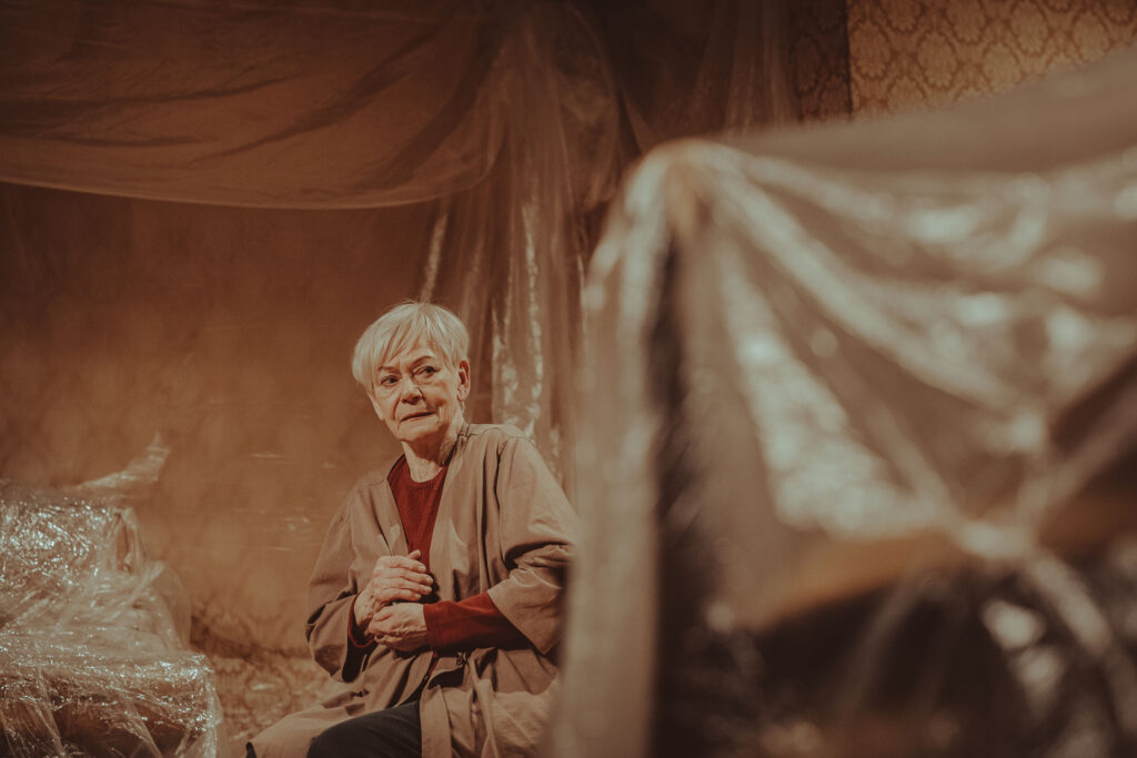 Zdjęcie ze spektaklu Wujaszek Wania. Starsza aktorka siedzi na środku sceny. Wokół są meble pokryte folią.