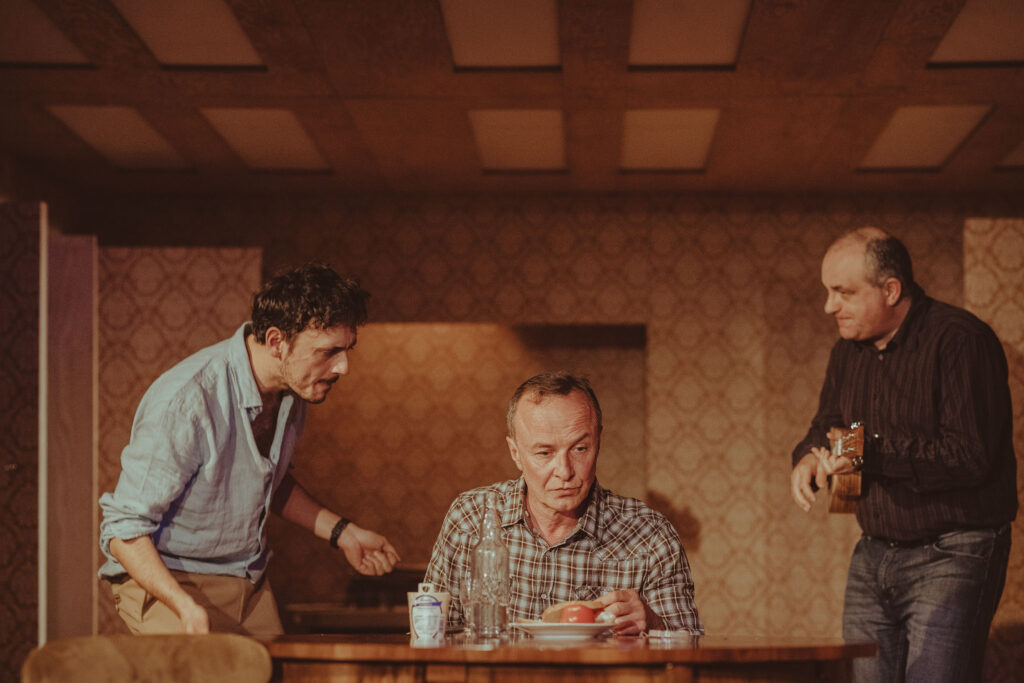 Zdjęcie ze spektaklu Wujaszek Wania. Aktor siedzi przy stole. Na stole jest talerz z chlebem i pomidorem, biały ser i szklana butelka. Z lewej strony stoi pochylony aktor. Z prawej strony aktor gra na ukulele. Na ścianie jest wzorzysta tapeta.