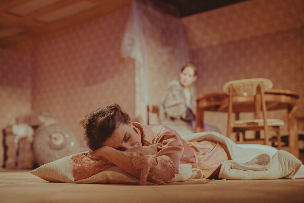 Zdjęcie ze spektaklu Wujaszek Wania. Aktorka leży na podłodze pod kołdrą, ma zamknięte oczy. Z tyłu przy stole siedzi druga aktorka.