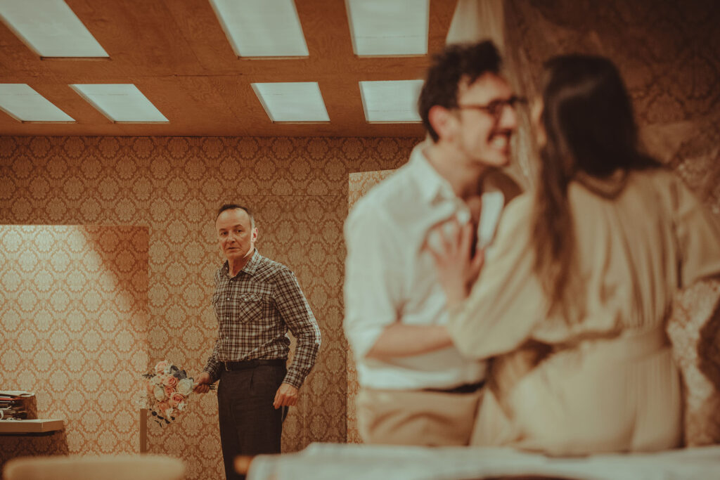 Zdjęcie ze spektaklu Wujaszek Wania. Aktor i aktorka stoją naprzeciwko siebie. Aktor się uśmiecha, a aktorka trzyma mu dłoń na piersi. Z tyłu przy drzwiach stoi aktor w średnim wieku z bukietem kwiatów w dłoni. Na ścianie jest wzorzysta tapeta.