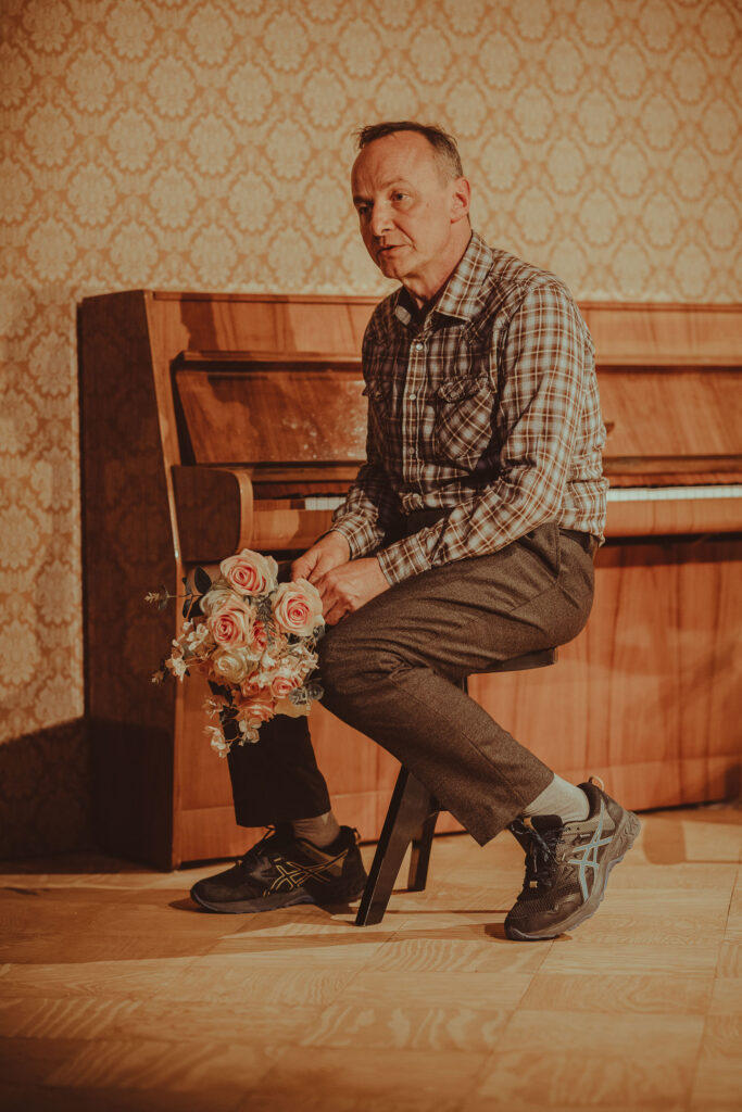 Zdjęcie ze spektaklu Wujaszek Wania. Aktor w średnim wieku siedzi bokiem na taborecie przy pianinie. Trzyma bukiet kwiatów. Na ścianie jest wzorzysta tapeta.
