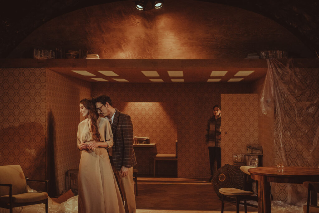 Zdjęcie ze spektaklu Wujaszek Wania. Na pierwszym planie aktor w marynarce stoi za aktorką w sukni. Stykają się głowami. Z tyłu w drzwiach stoi druga aktorka i patrzy na nich. W tle są meble, resztki folii i wzorzysta tapeta na ścianie.