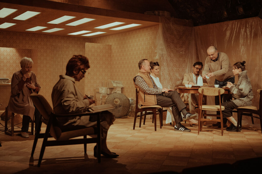 Zdjęcie ze spektaklu Wujaszek Wania. Przy stole siedzą aktorzy i aktorki, przed nimi talerze, kanapki, filiżanki, dzbanek. Obok na krzesłach dwie starsze aktorki, jedna trzyma książkę, druga je chleb. W tle meble, folia i wzorzysta tapeta.