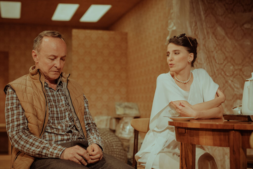 Zdjęcie ze spektaklu Wujaszek Wania. Młoda aktorka i aktor w średnim wieku siedzą przy stole. On spuszcza wzrok, ma koszulę w kratę i serdak. Ona patrzy na niego, jest elegancka, ma perły, okulary na włosach. W tle meble, folia i wzorzysta tapeta.