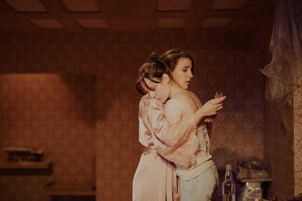 Zdjęcie ze spektaklu Wujaszek Wania. Dwie młode aktorki przytulają się. Jedna trzyma w dłoni mały kieliszek. W tle są resztki folii i wzorzysta tapeta na ścianie.