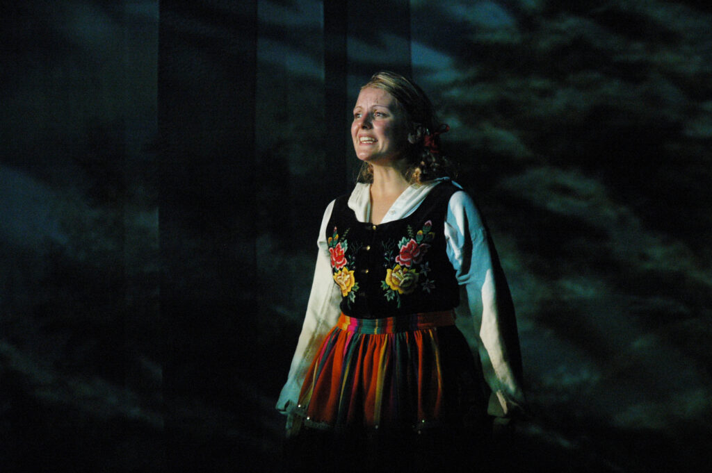 Zdjęcie ze spektaklu Pyza na polskich dróżkach. Zbliżenie. Aktorka z warkoczami w łowickim stroju ludowym. Aktorka otwiera usta, jest przejęta.