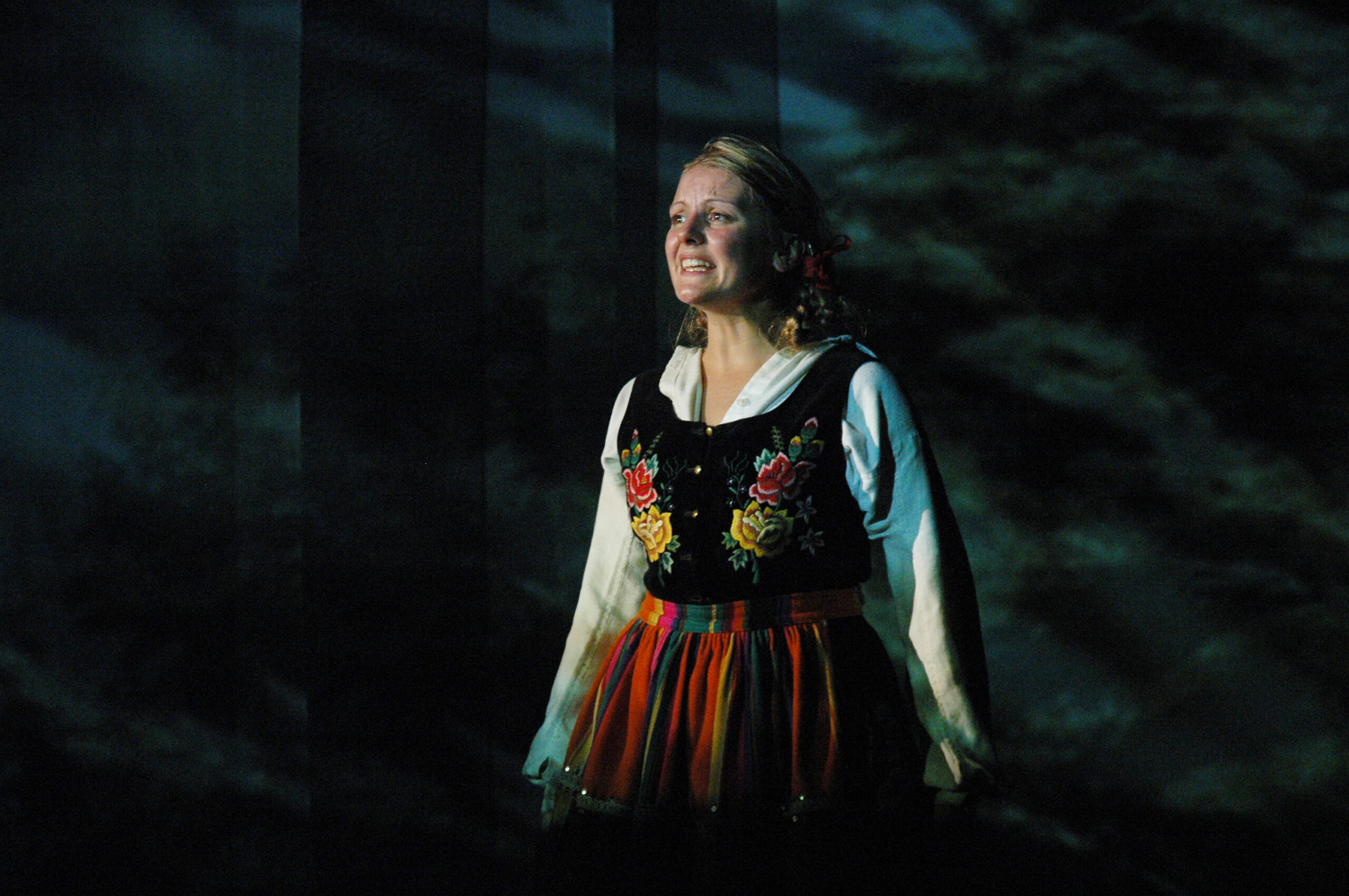 Zdjęcie ze spektaklu Pyza na polskich dróżkach. Zbliżenie. Aktorka z warkoczami w łowickim stroju ludowym. Aktorka otwiera usta, jest przejęta.