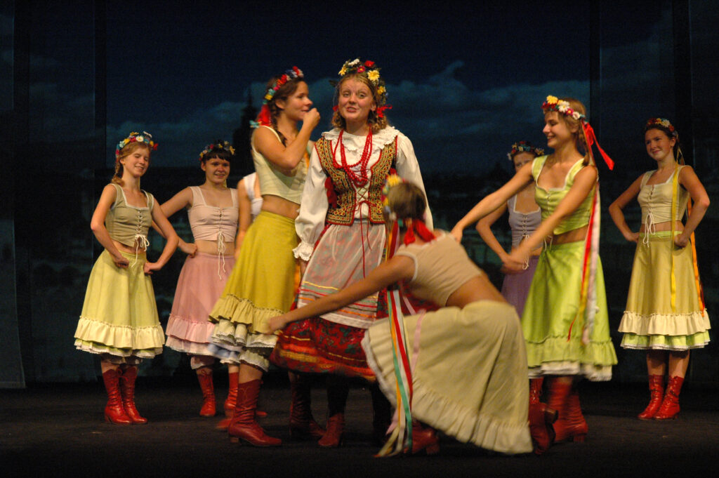 Zdjęcie ze spektaklu Pyza na polskich dróżkach. Scena zbiorowa. Na środku stoi aktorka z warkoczami w krakowskim stroju ludowym. Na głowie ma wianek. Wokół niej tańczą dziewczyny w spódnicach, czerwonych trzewikach i wiankach na głowach.