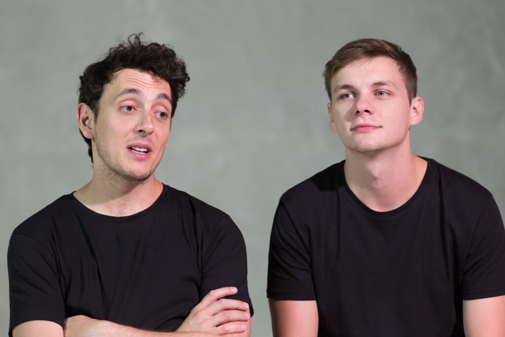 djęcie ze spektaklu Trik Patryka. Zbliżenie. Dwaj aktorzy. Aktor po lewej stronie ma ręce skrzyżowane na piersiach. Aktorzy patrzą w różne strony.