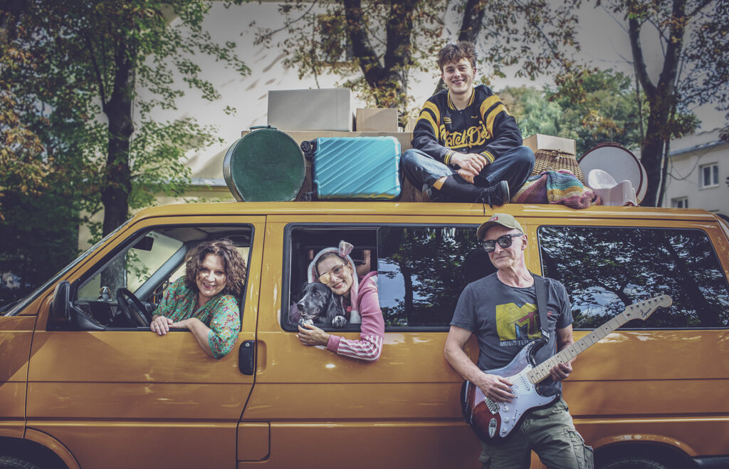 Zdjęcie ze spektaklu Preskot. Aktorka siedzi za kierownicą vana i wychyla się przez okno. Przez drugie okno wychyla się młoda aktorka z psem. Na dachu vana siedzi młody aktor z walizkami. Aktor z gitarą elektryczną opiera się o vana.